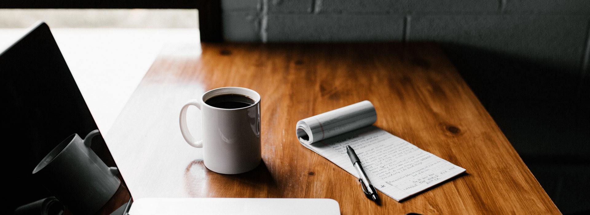 Notepad with writing, a pen, a laptop, and a black coffee in a white mug sit on a wooden desk tabletop